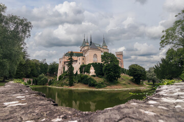 Fototapeta premium Ground-level view of Bojnice Castle across the water, framed by stone foreground, trees and a dramatic cloudy sky in Bojnice, Slovakia.