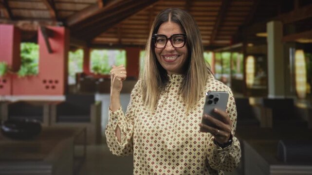Woman holding smartphone giving thumbs up in building lobby wearing patterned blouse and glasses; warm connection joy.