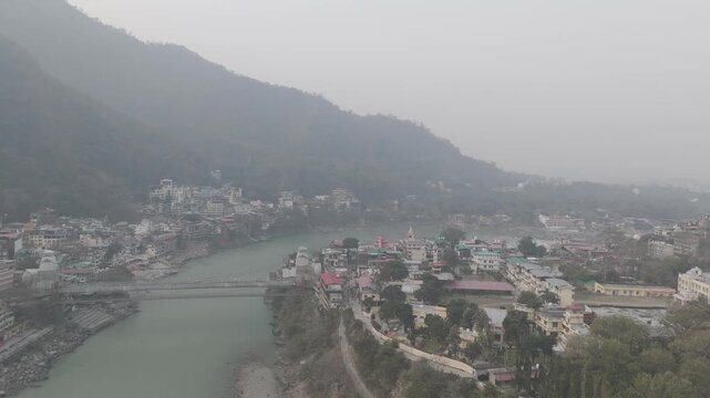Aerial view of Lakshman Jhula in Rishikesh, Uttarakhand, with the Ganga River flowing below, and temples, houses, and buildings along the riverbank against misty mountains on a winter morning.