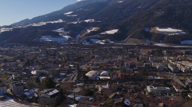 Aerial view of Brixen Bressanone, South Tyrol shows sports domes, railway, clustered roofs, terraced fields, and snow patches as the camera slowly sweeps in daylight.