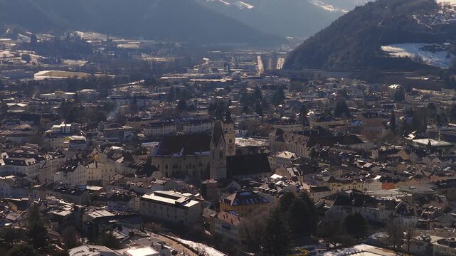 Aerial sweep over Brixen, South Tyrol, in the Eisack Valley, with twin steeple church, tiled roofs, narrow streets, snow patches, and a highway in late winter light.