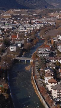 Aerial view of Brixen, Italy shows the Eisack River winding past bridges and a church steeple, red tiled houses, apartments, industry, vineyards, and orchards.