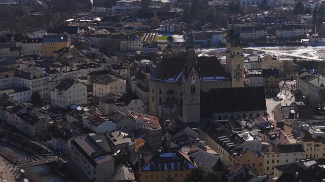 Aerial view of Brixen, South Tyrol, shows the duomo, clock tower, pastel roofs, the Isarco river, snow patches, and light pedestrian activity in late winter.