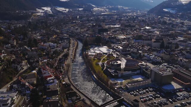 Aerial view of Brixen in South Tyrol shows the Eisack River, the cathedral, a circular solar roof plant with steam, cars moving, snow patches, and winter daylight.