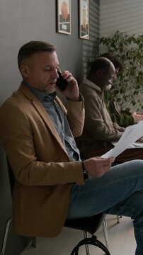 Vertical shot of middle-aged man in blazer speaking on phone, holding documents in modern bank waiting area