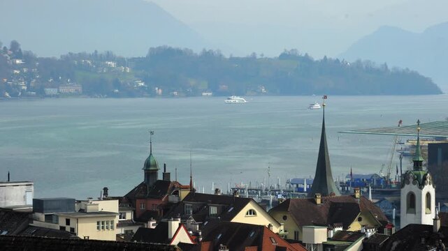 Static wide elevated shot over the historic rooftops and church towers of Lucerne with several passenger ships on the lake and mountains in the distance