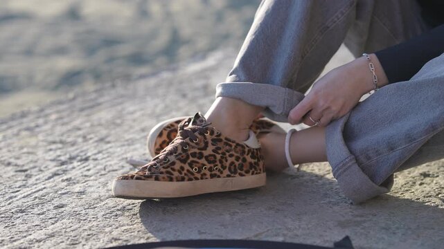 Close-up view of a young woman's hands rolling up the cuffs of her grey jeans. She is sitting outdoors on a concrete surface wearing stylish leopard print sneakers