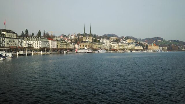 LUCERNE, SWITZERLAND - MARCH 06 2026: Panning shot across the scenic waterfront of Lake Lucerne featuring grand hotels and the historic Hofkirche St. Leodegar twin towers under a clear sky