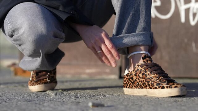 Close-up of a stylish woman crouching down to roll up the cuffs of her gray jeans, showing off her trendy leopard print sneakers and anklet
