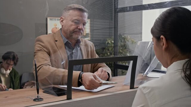 Middle-aged male customer handing cash money to bank teller across glass counter in modern bank office