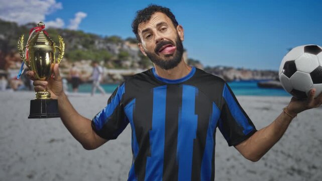 Man in blue jersey holding trophy and soccer ball, shrugs with a smirk and beard visible on a studio beach set; pride.
