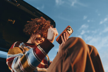 Smiling woman with curly hair wearing a rainbow sweater sitting in a car trunk holding a smartphone, enjoying a sunny day with clear blue sky background, lifestyle and happiness concept. © SHOTPRIME STUDIO