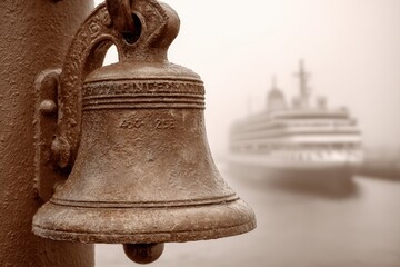 Obraz premium A weathered bronze ship bell with intricate detailing, contrasting with the modern cruise ship in the hazy background, creating a sense of history and maritime travel.