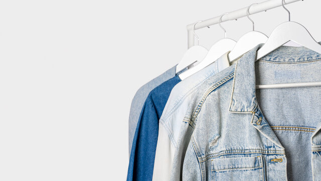 Denim jackets, blue shirts on white wooden hangers, clothing rack on light gray background. Concept of casual fashion wardrobe, modern denim collection and everyday clothing style. Copy space, banner