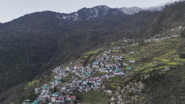 Aerial view of Triyuginarayan village in the Garhwal Himalayas, showing colorful houses, terrace farming slopes, and forests of pine, oak, rhododendron, and deodar with snow-capped peaks.