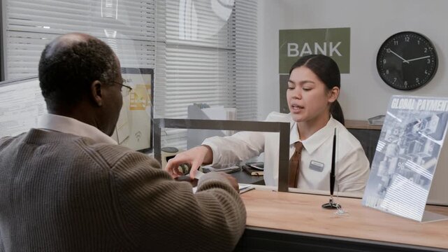 Elderly African American customer wearing glasses and sweater talking to bank teller in white shirt through glass counter