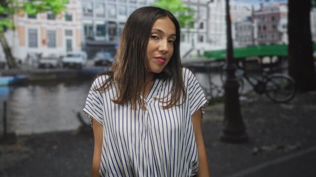 Hispanic woman tilts head with slight smirk wearing striped blouse on canal street in amsterdam; playful curiosity.