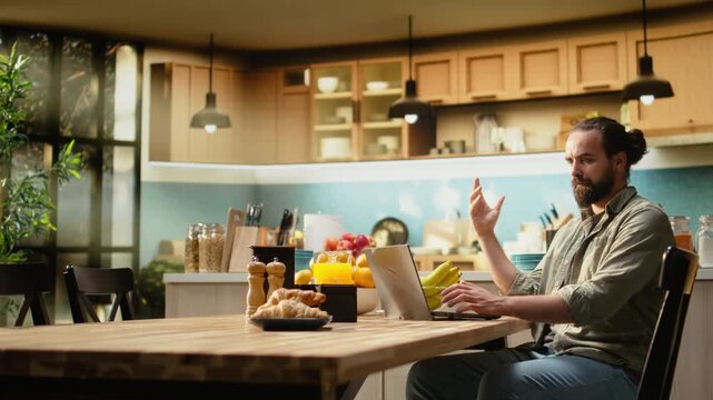 Disoriented man being confused and uncertain with his work at laptop, working from home in the kitchen. Male freelancer unsure frowning at the screen, ambiguous teleworking.