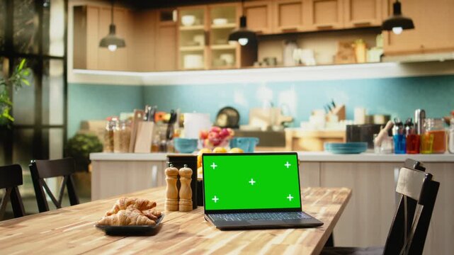 Green screen laptop on wooden table in empty bright kitchen, breakfast prepared with pastry, croissants and coffee. Warm tones on rustic furniture and kitchenware, cozy decorative items.