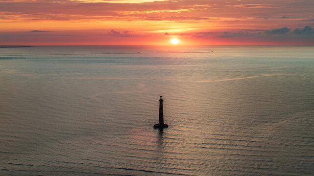 Panormic aerial view of ocean and Morris Island Lighthouse in Folly Beach near Charleston, South Carolina during morning sunrise above horizon over calm waters and golden skies landscape seascape