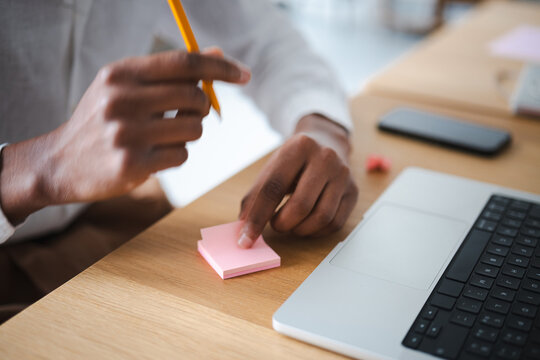 A person's hands are shown at a desk, one holding a pencil and the other touching a stack of pink sticky notes next to a laptop.