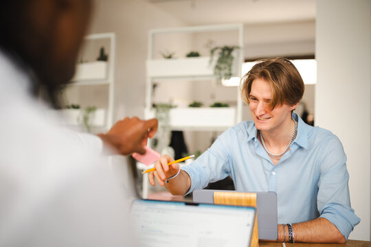 Two colleagues collaborate on a project, one handing a pink sticky note to the other who holds a pencil, both focused on their work.