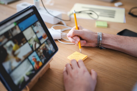 A person's hand holds a pencil, writing on a sticky note that says "Investment Business Profits" while looking at a tablet.