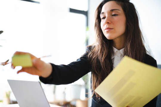 A focused businesswoman in a suit holds a document and a sticky note while working at her desk with a laptop.