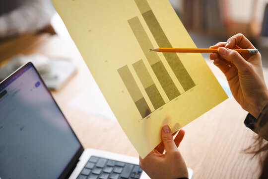 A person holds a bar graph with a pencil, reviewing data next to a laptop.