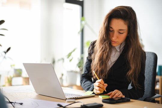 A focused woman in a suit works at her desk, writing on a notepad next to her laptop and phone.