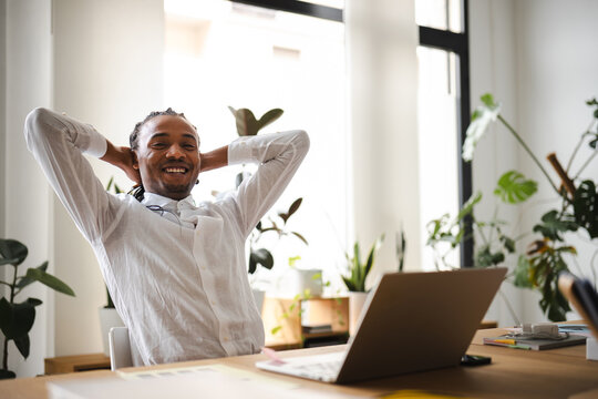 A man in a white shirt smiles with his hands behind his head, relaxing at his desk with a laptop and plants in the background.