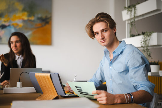 A young man in a blue shirt reviews documents at a desk, with a colleague in the background working on a laptop.