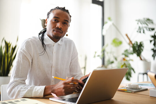 A man with braided hair sits at a desk, holding a pencil and looking at a laptop. He is in a bright, modern office with plants in the background.