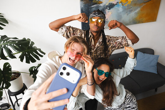 Three friends pose for a selfie, two wearing heart-shaped sunglasses and one flexing his muscles, capturing a moment of joy and connection.