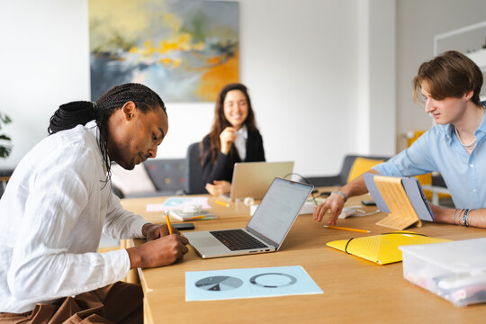 A diverse group of young professionals collaborate around a table, engaged in work with laptops and tablets, fostering a productive and modern office environment.