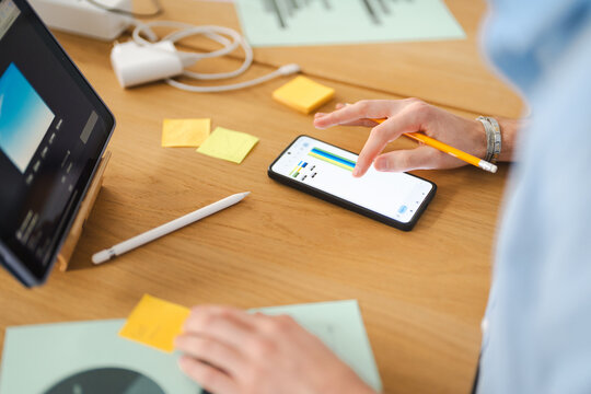 A person's hands interact with a smartphone displaying a graph and a tablet showing an image, surrounded by sticky notes and a pencil on a wooden desk.