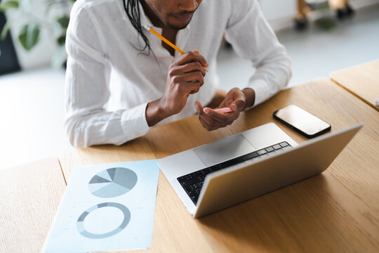 A man in a white shirt holds a pencil and a sticky note while working at a desk with a laptop and charts.