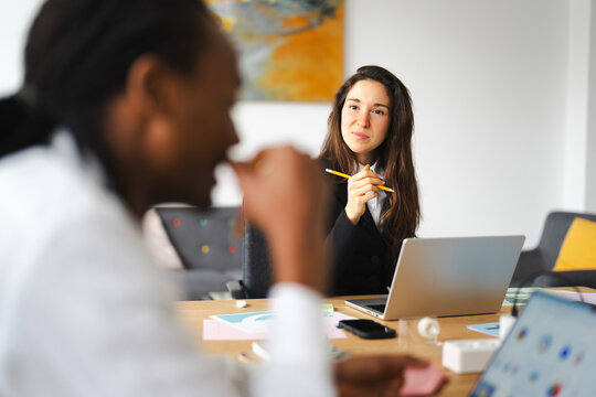 Two women in a modern office setting collaborate on a project, one holding a pencil and looking thoughtfully at the other.