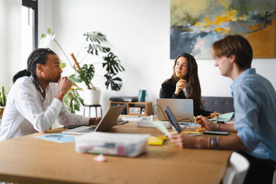 Three colleagues collaborate around a table, engaged in discussion and working on laptops and papers in a bright, modern office space.