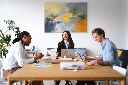 A diverse team collaborates around a table, engaged in discussion and working on projects. They are in a modern office setting with artwork on the wall.