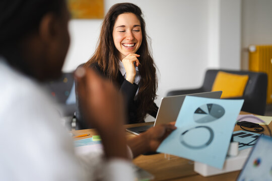 Two colleagues discuss charts and data during a meeting, one woman laughs heartily while the other points to a document.