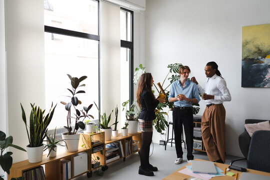 Three colleagues in a bright, plant-filled office share a moment of conversation and drinks, fostering a positive work environment.