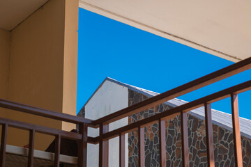 Naklejka premium View from a balcony featuring brown railings, a stone-patterned building, and a vibrant blue sky. Clean architectural lines and high contrast create a minimalist urban scene.