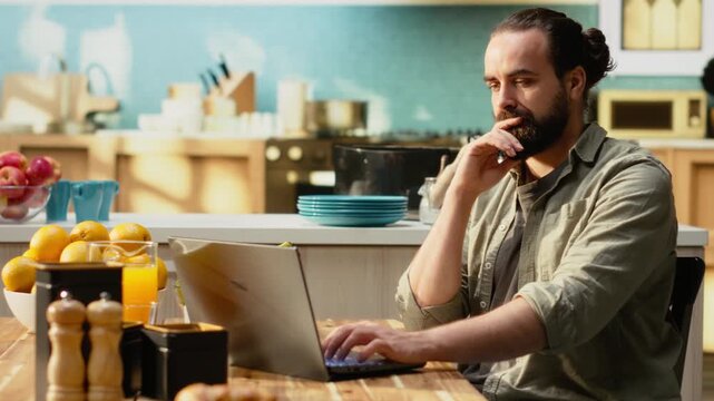 Adult working remotely from his home setup in the kitchen, focused on solving daily administrative tasks using a laptop and wireless internet. Digital nomad lifestyle, flexible work routine.