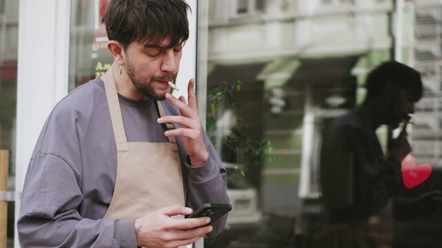 Barista takes a break while smoking outside a cafe and checking messages on a smartphone during working hours