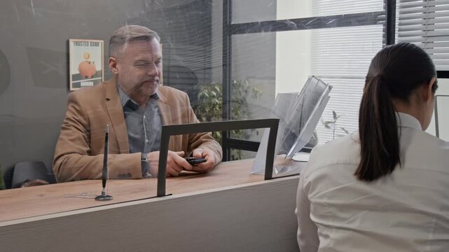 Middle-aged male customer handing documents to bank teller through transparent divider at local bank office