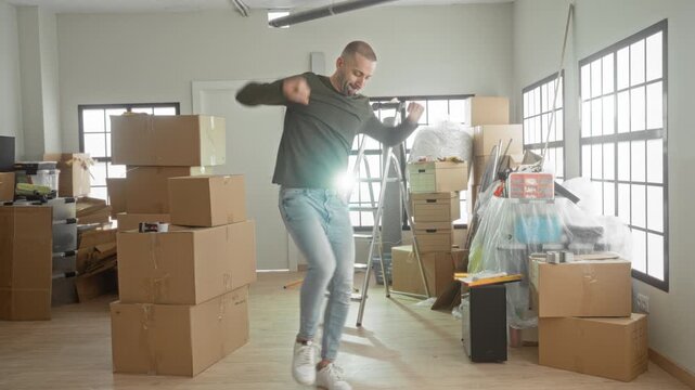 Man midair jumping beside stacked cardboard boxes and a ladder in a cluttered room of a building during moving day; joy moving celebration.