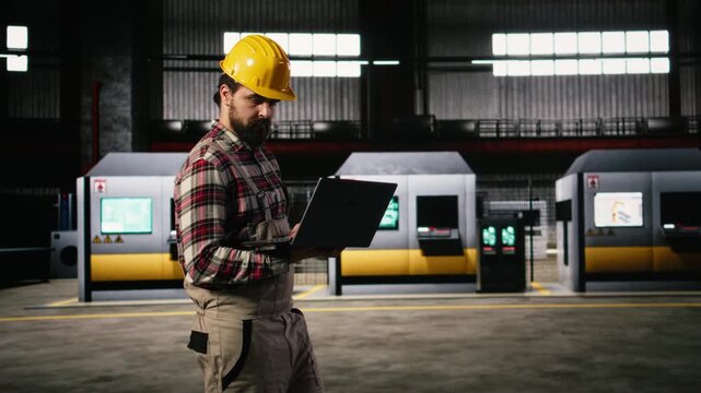 Professional engineer walking through heavy industry workshop in factory, reviewing equipment and metalwork quality on his laptop. Industrial engineering plant features manufacturing production.