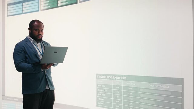Manager analyzes revenue, expenses and net profit displayed on presentation wall using laptop. African american man uses notebook to review quarterly KPIs results, camera A