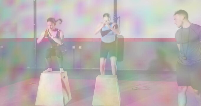 Two women adopting stance and performing box jumps on plyo boxes in gym, trainer supervising form